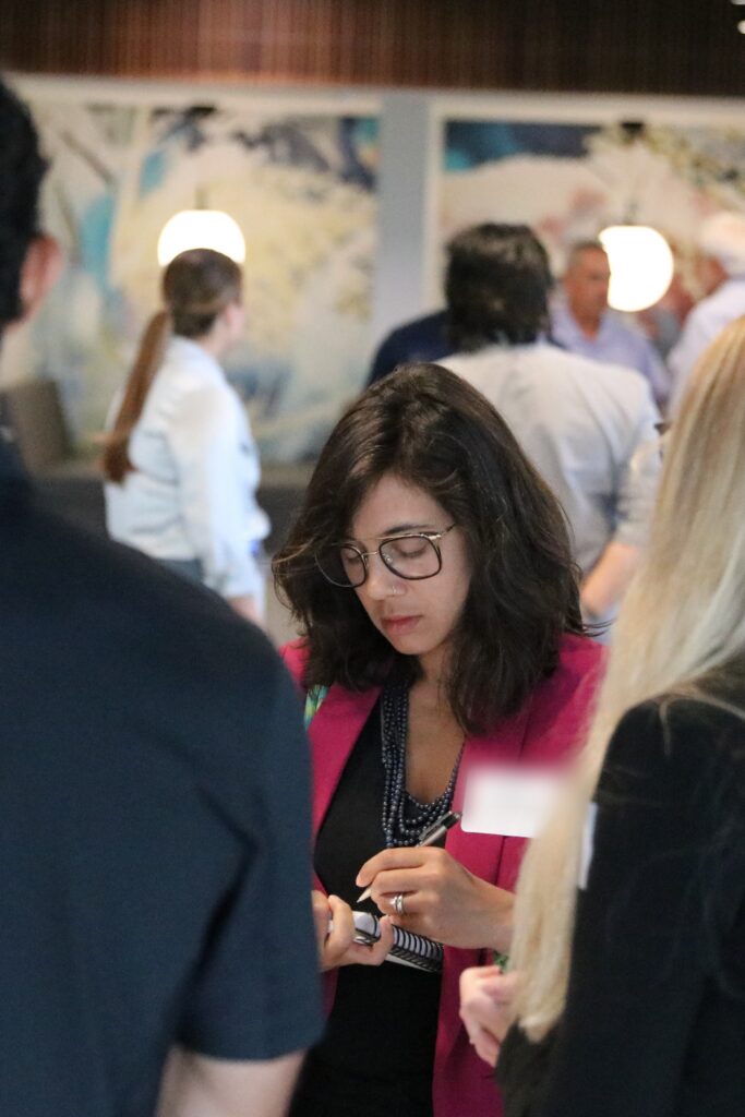Person in glasses and a pink blazer taking notes while talking with colleagues at a networking event
